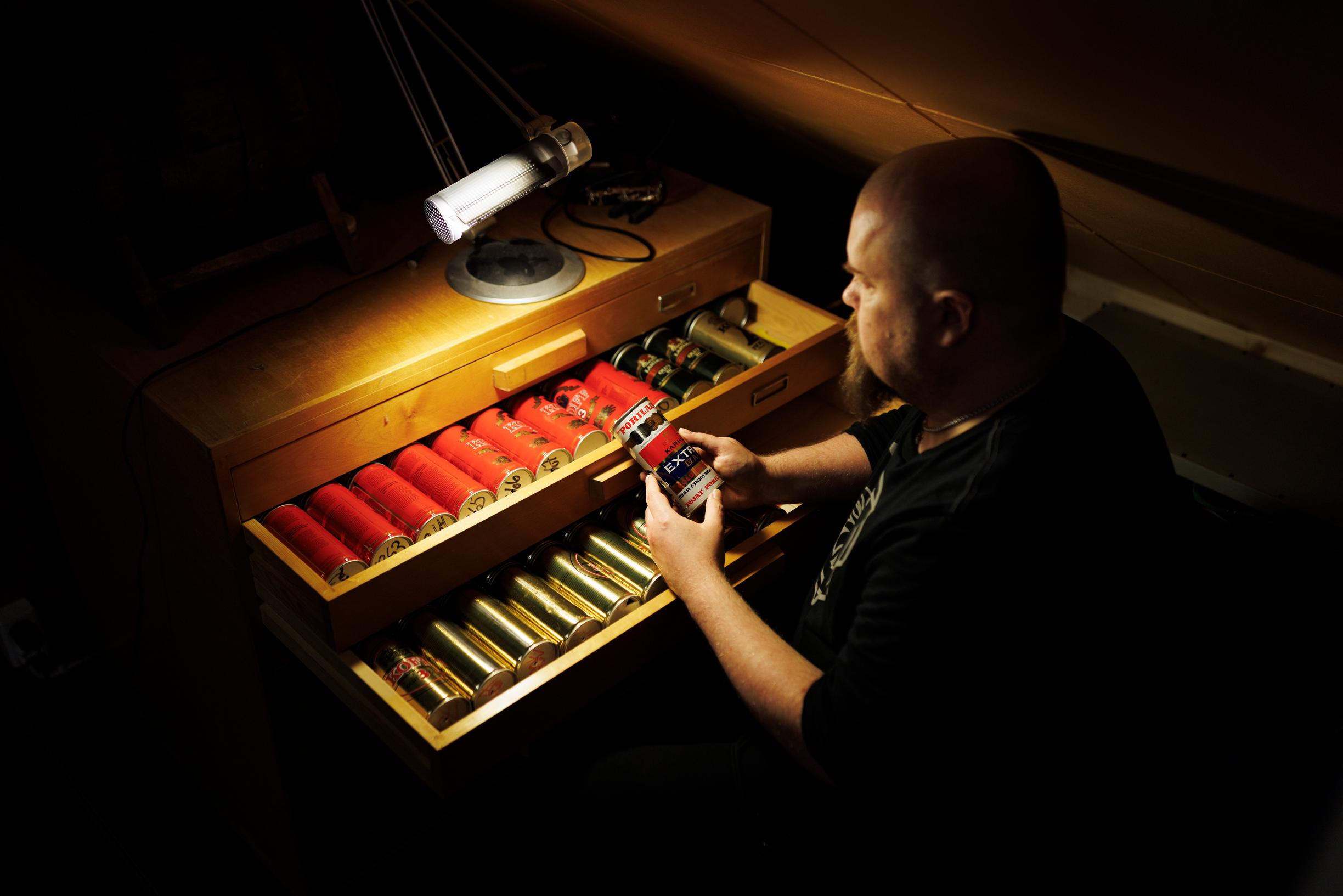 Tummasävyinen kuva, jossa lipaston vetolaatikossa rivi kiiltäväpintaisia oluttölkkejä. Lipaston äärellä mies istumassa kädessään yksi tölkki.